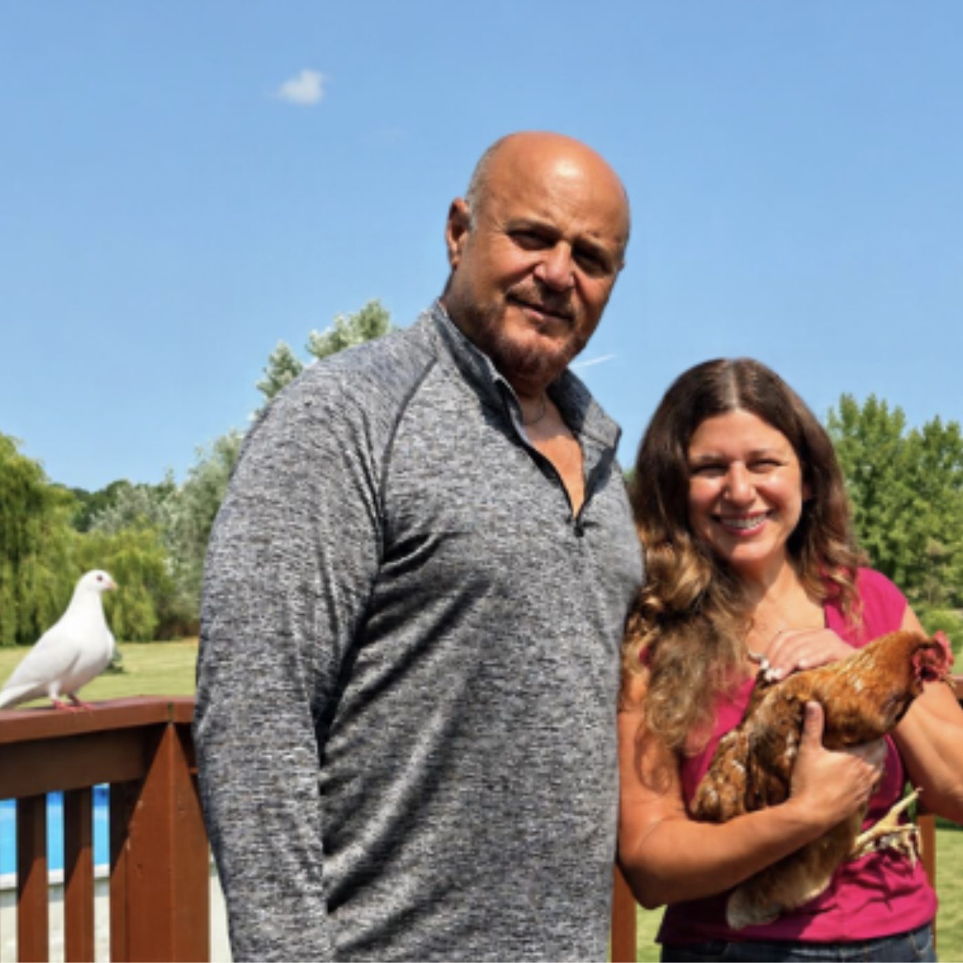 Brenda and Asim smiling on the deck at AB Healing, Brenda holding one of their chickens while a white dove perches nearby